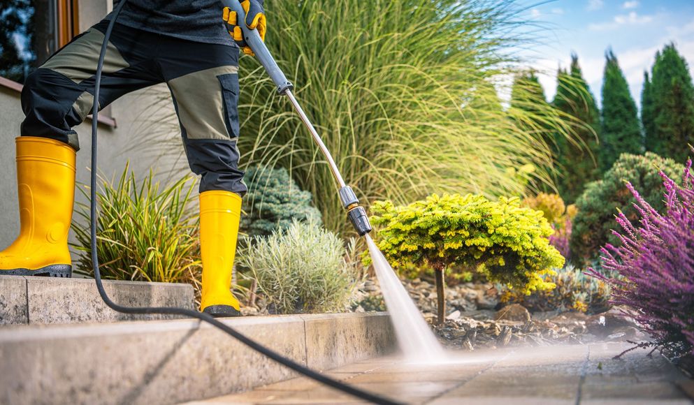 A person in yellow boots using a pressure washer to clean a paved walkway in a garden setting.