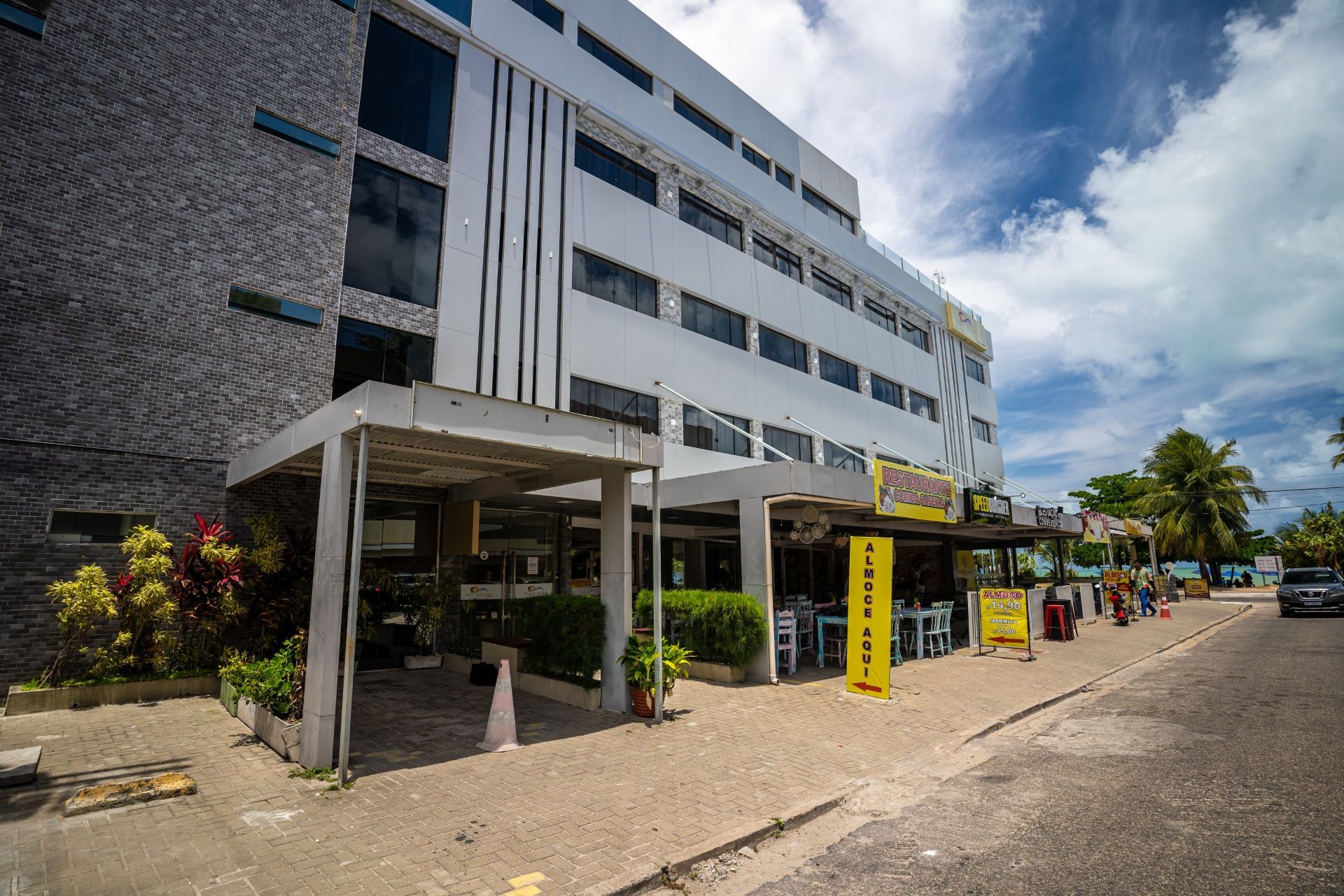 Exterior de um prédio branco de vários andares com entrada coberta e carros estacionados em um dia ensolarado.