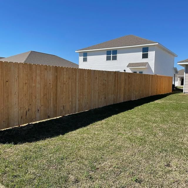 A wooden fence surrounds a grassy backyard with a white house and blue sky in the background.