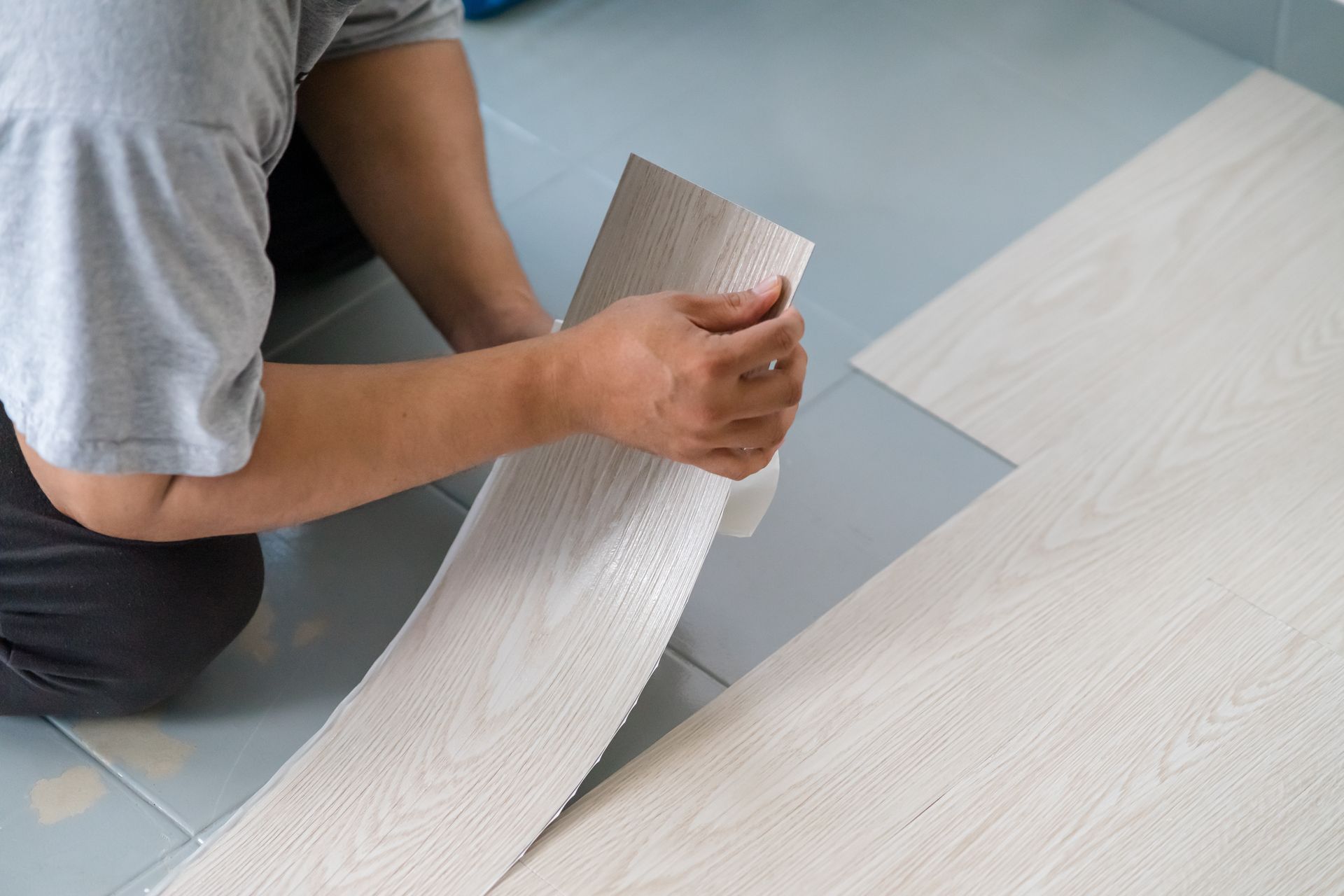 Person installing light-colored vinyl flooring; hands hold and arrange a plank on a blue-tiled floor.