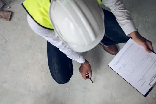 A man wearing a hard hat is kneeling down and writing on a clipboard.