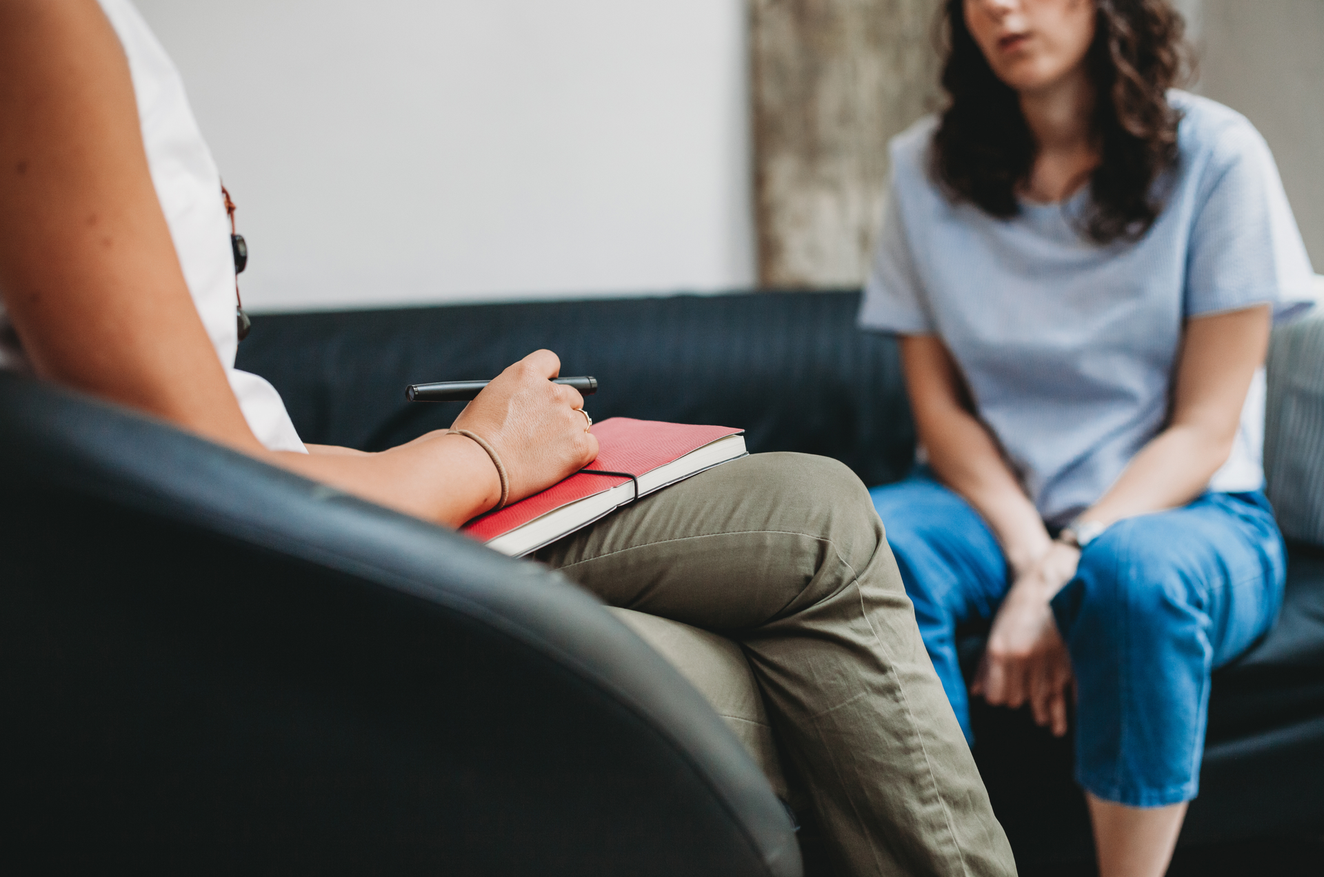 Therapist and patient in session, seated on a black couch. Therapist holds a notebook. Patient looks on.