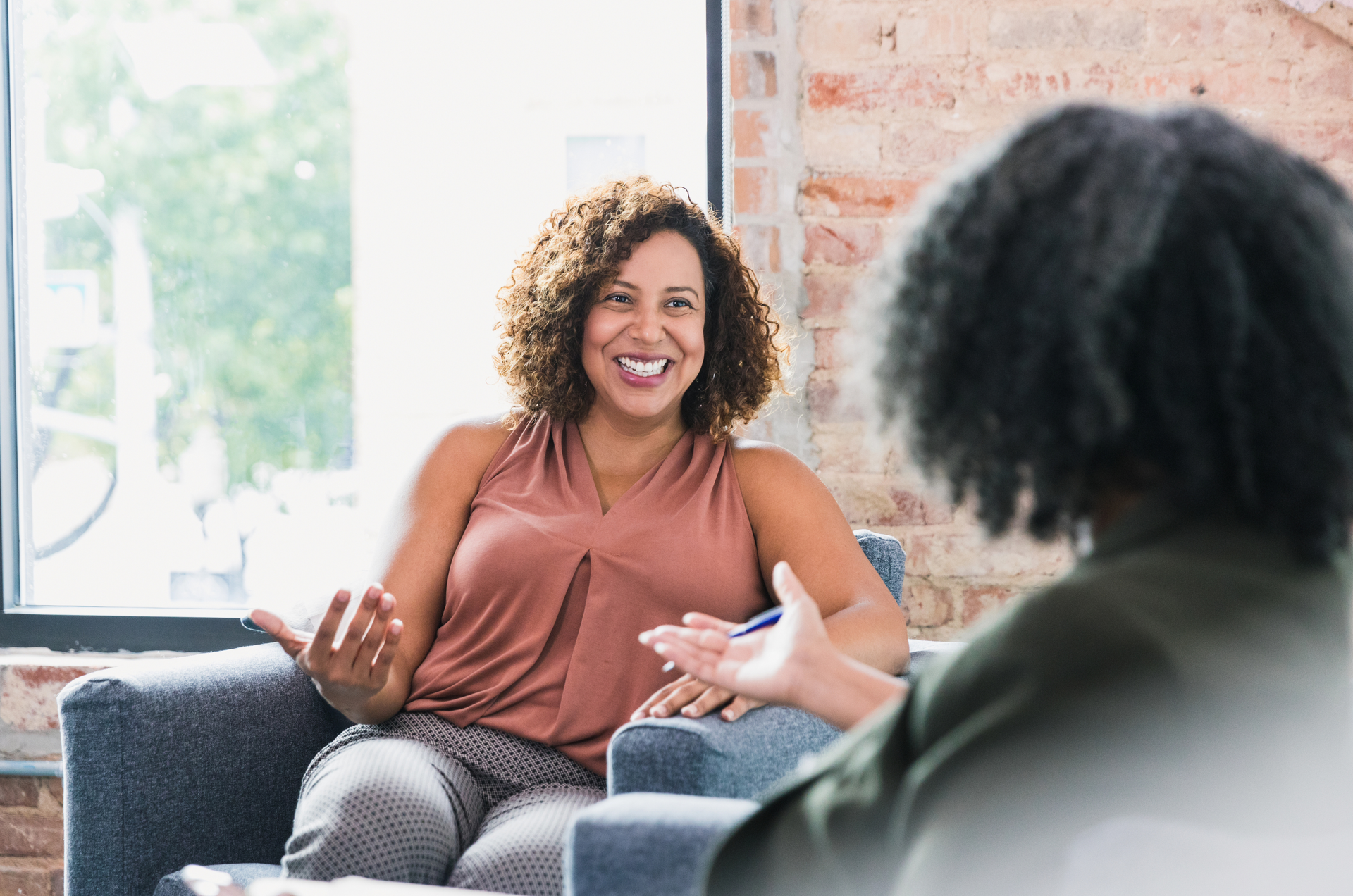Woman in peach top smiles, gesturing in conversation with another in a counseling session.