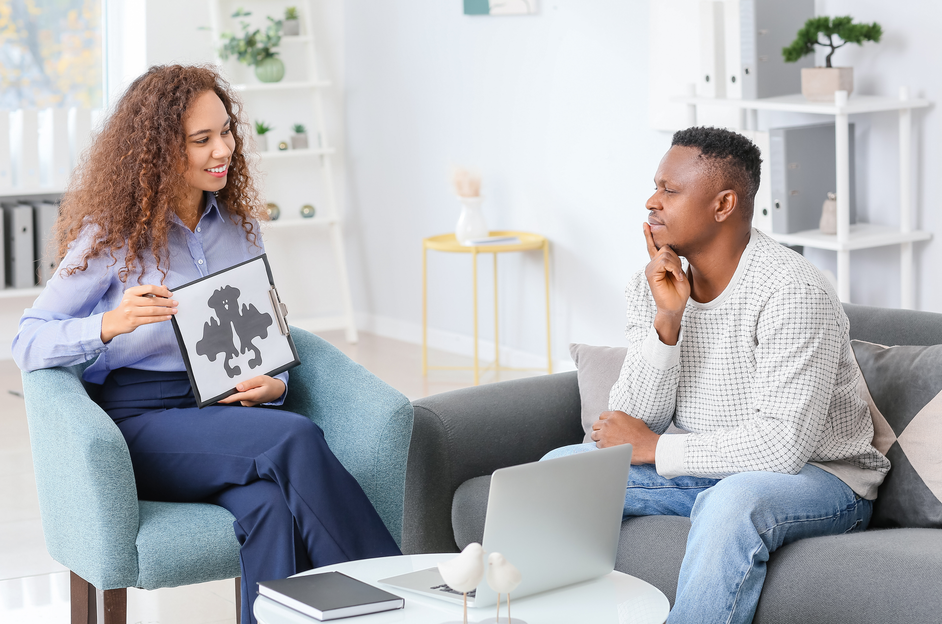 Therapist holding inkblot card speaks with a patient in a living room setting; laptop present.