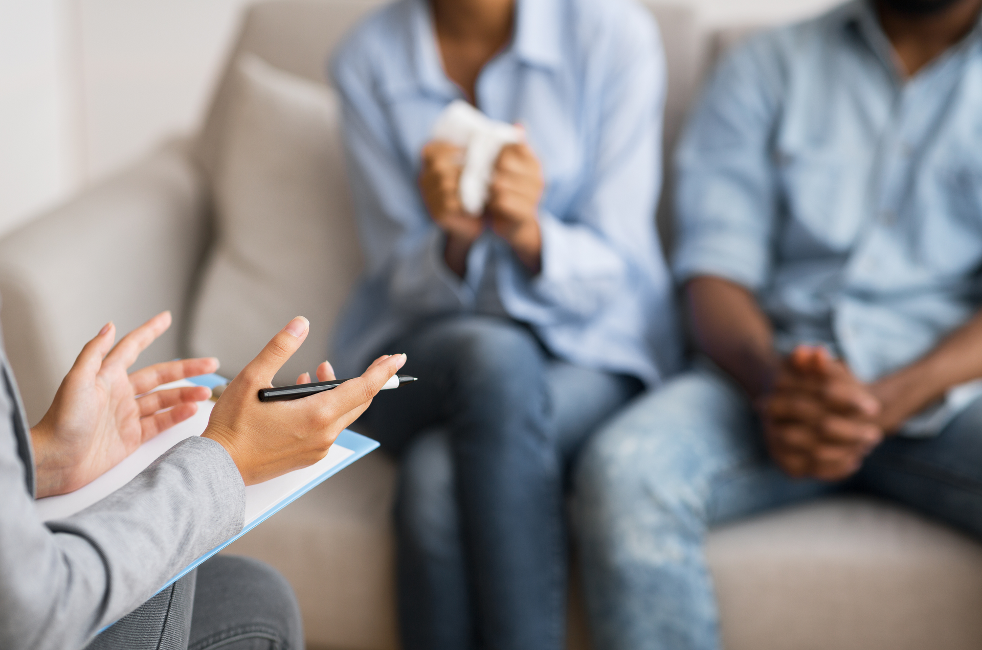 Therapist consults with a couple on a sofa. One person holds tissue; the other has hands clasped.