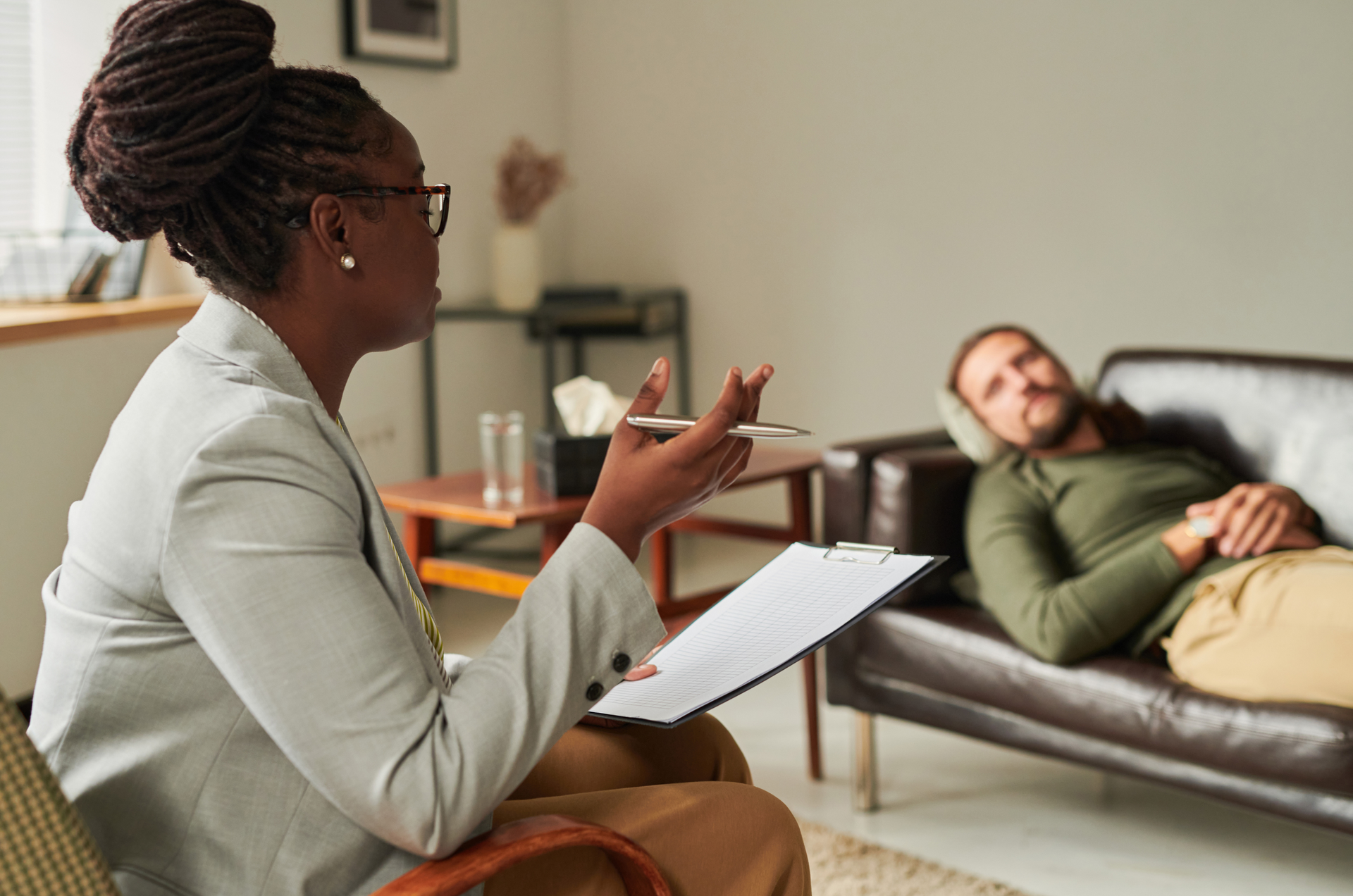 Therapist in light suit speaks to client lying on couch in an office, holding a notepad.