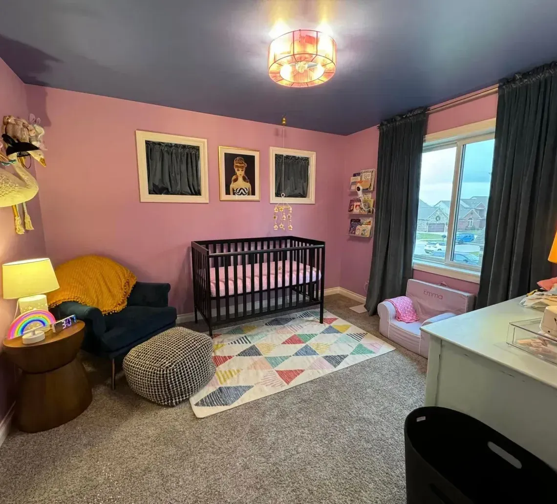 A nursery with lavender walls, a dark ceiling, a crib, a blue armchair with a yellow blanket, and a geometric rug.