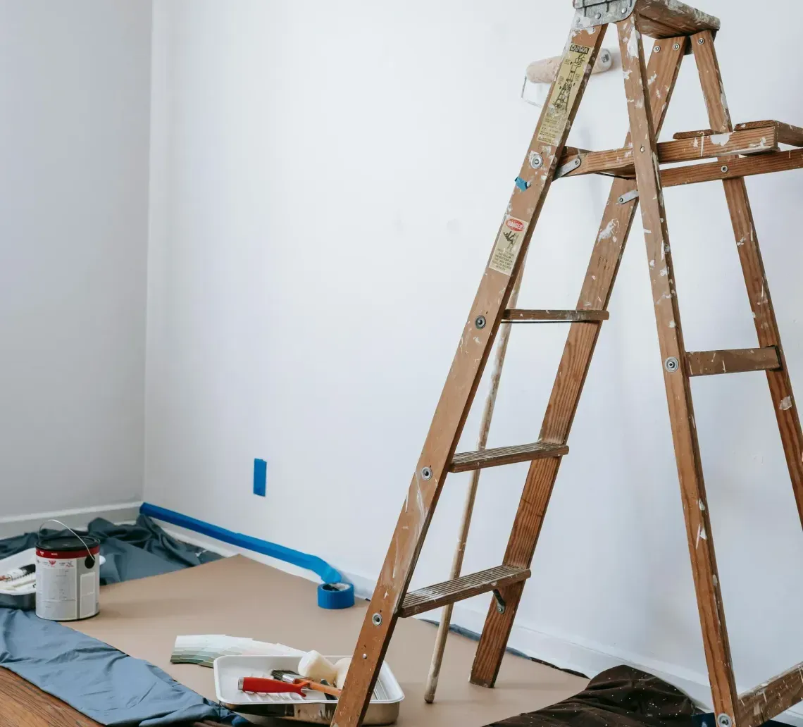 A wooden ladder stands on a drop cloth in a room prepared for painting, with painting supplies and floor tape visible.