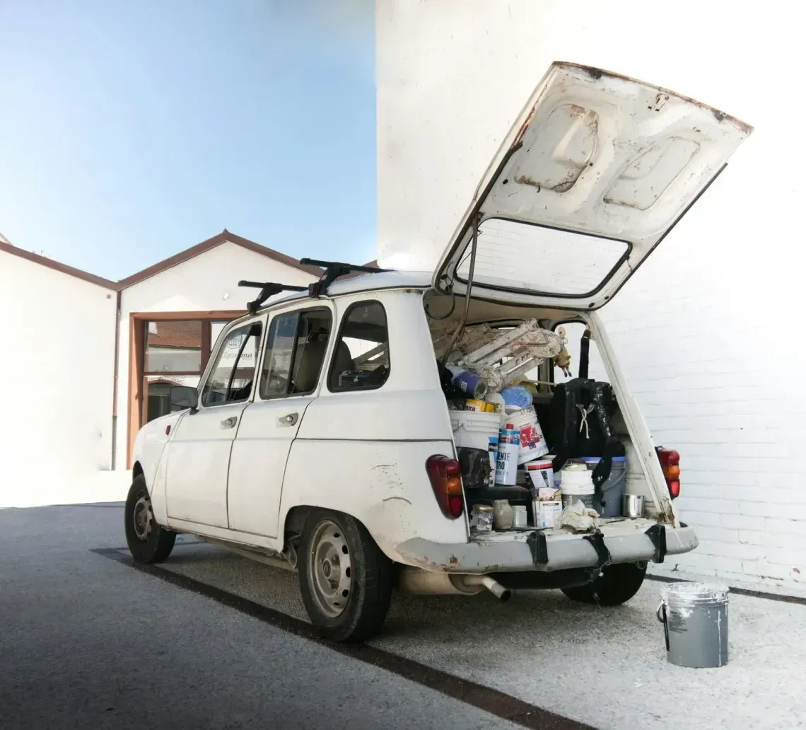 A white hatchback parked by a white wall with its trunk open, packed with various items and belongings.