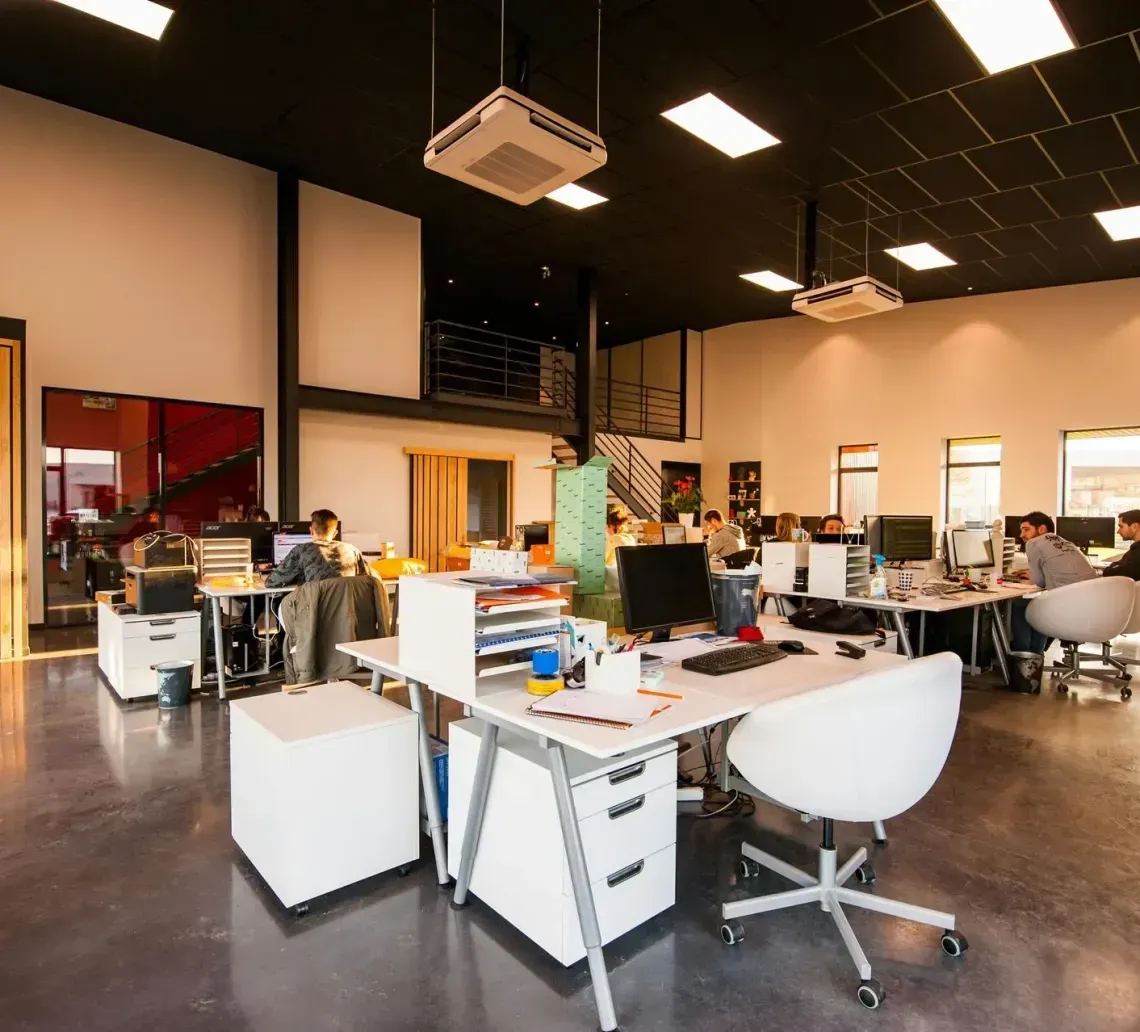 A modern, open-plan office with white desks, computers, and employees working, set against a dark ceiling and polished floor.