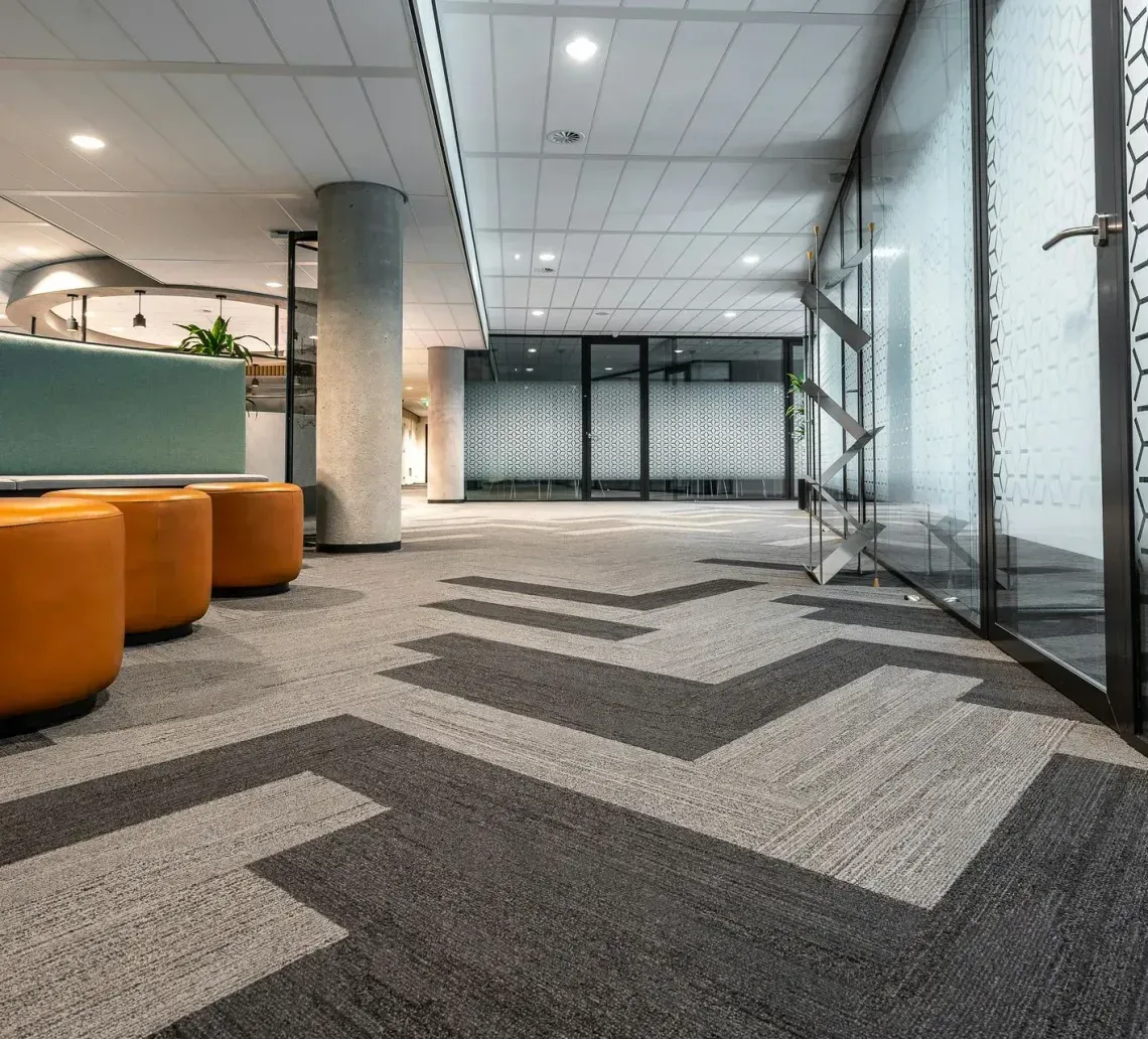 A modern office interior featuring three orange poufs, geometric grey carpet, a support pillar, and glass-partitioned rooms.
