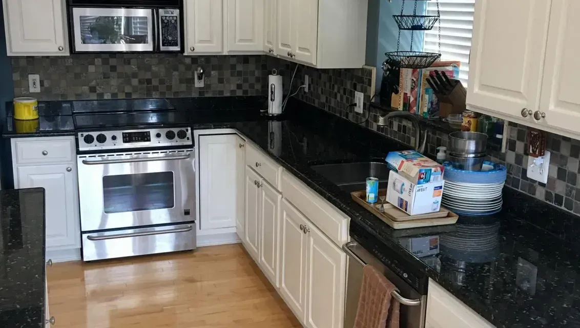 A modern kitchen with white cabinets, black granite countertops, stainless steel appliances, and light wood flooring.