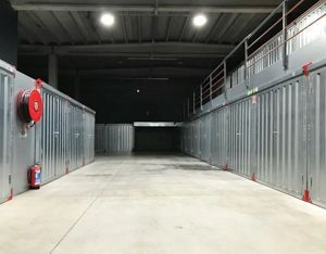An indoor corridor of a self-storage facility featuring metal storage units, concrete flooring, and overhead lighting.