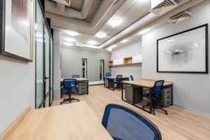 Modern office space with light wood desks, blue mesh chairs, and large framed artwork on white walls.