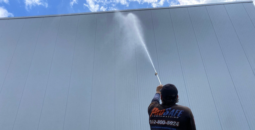 Person power washing a white building's wall with a long sprayer, against a partly cloudy blue sky.
