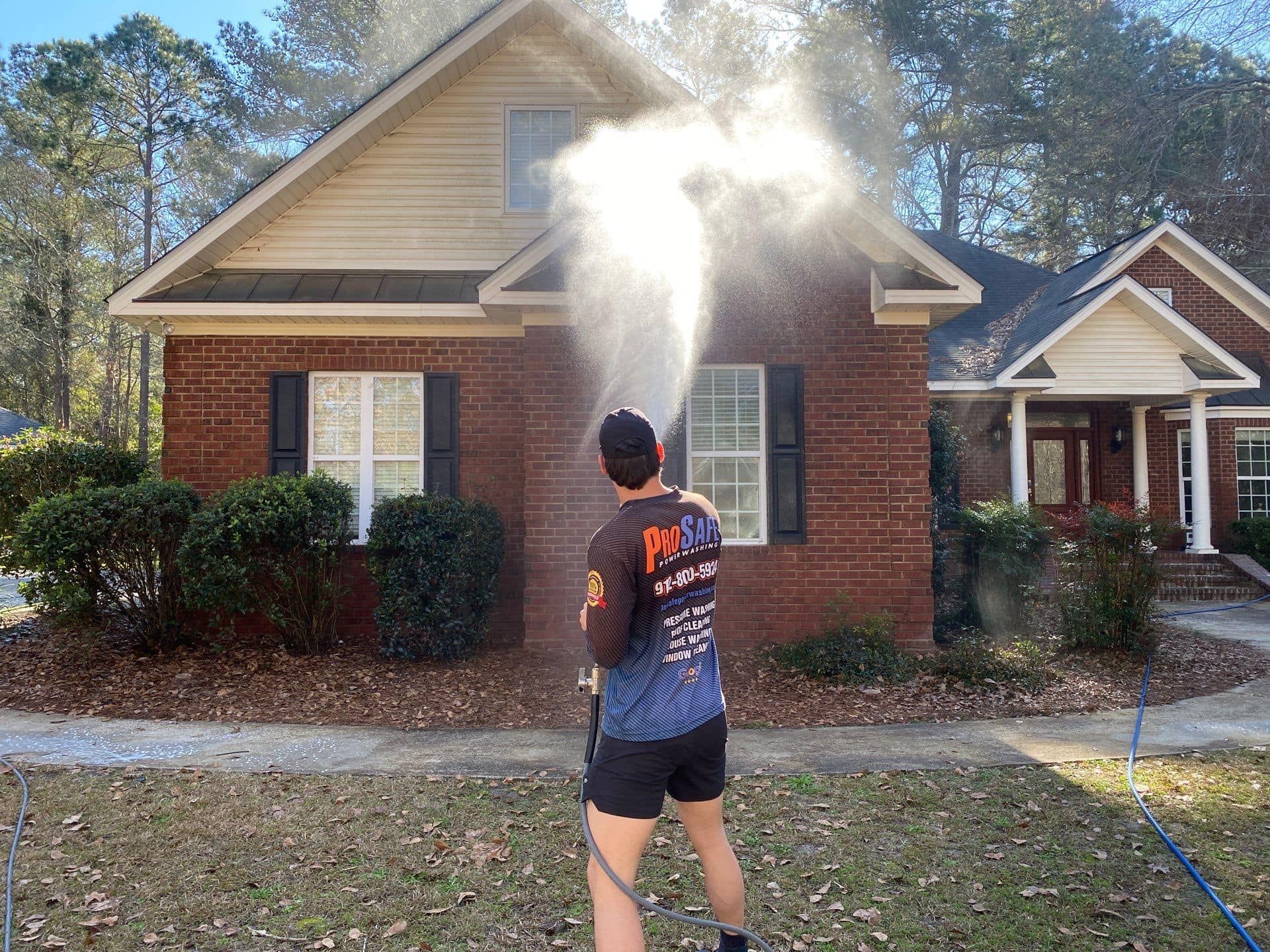 A man is spraying water on a brick house with a hose.