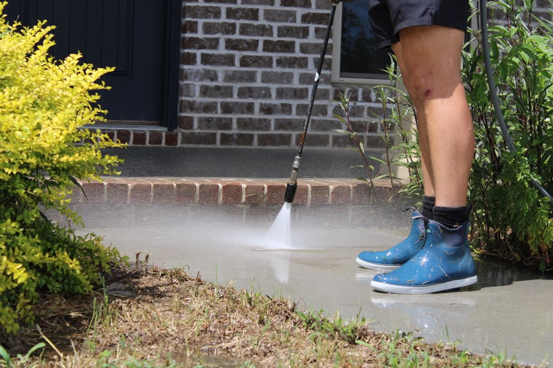 Person power washing a concrete sidewalk in front of a brick house.