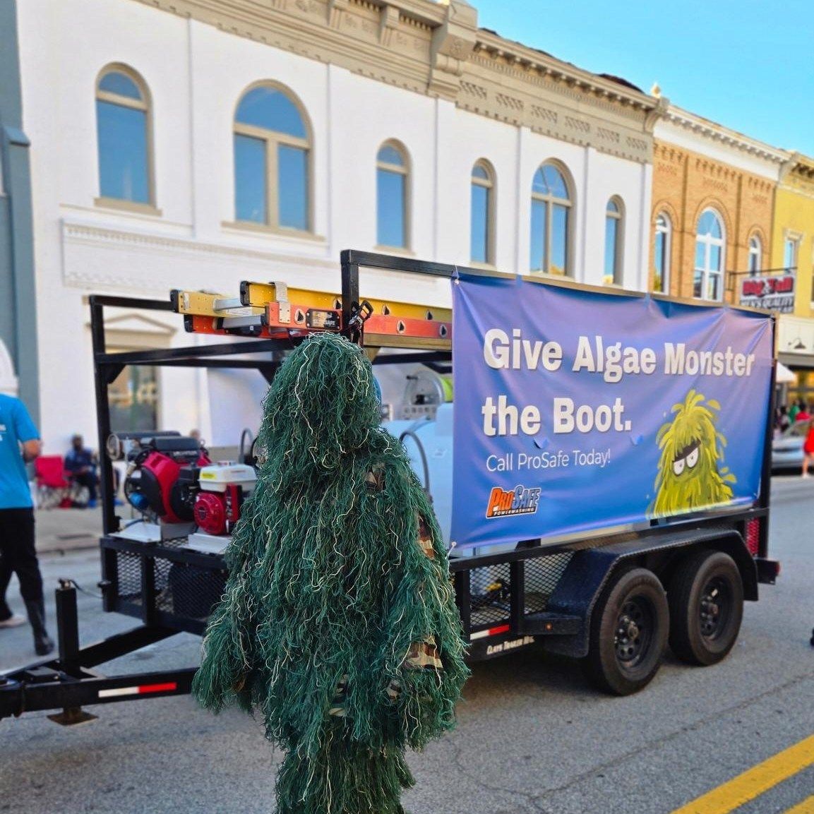 A person in a green ghillie suit stands next to a utility trailer with a banner reading