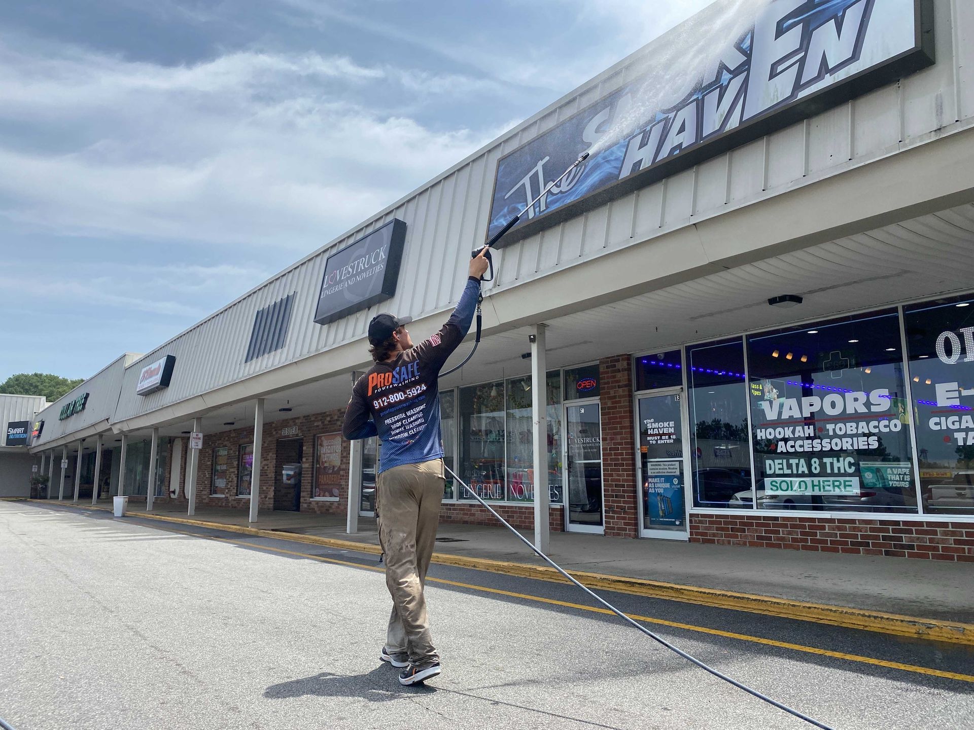 A person in a blue and black uniform uses a power washer to clean the sign of a shop labeled 
