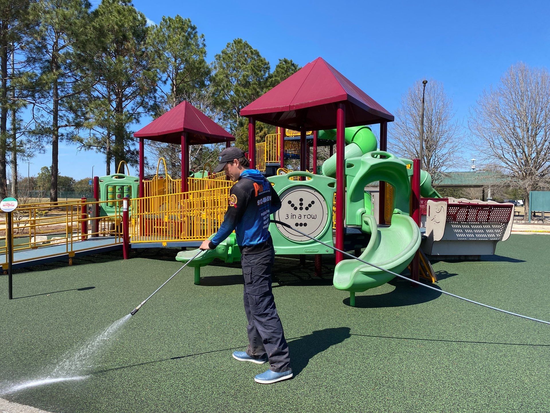 A maintenance worker pressure washes the rubber safety flooring of a sunny outdoor playground.