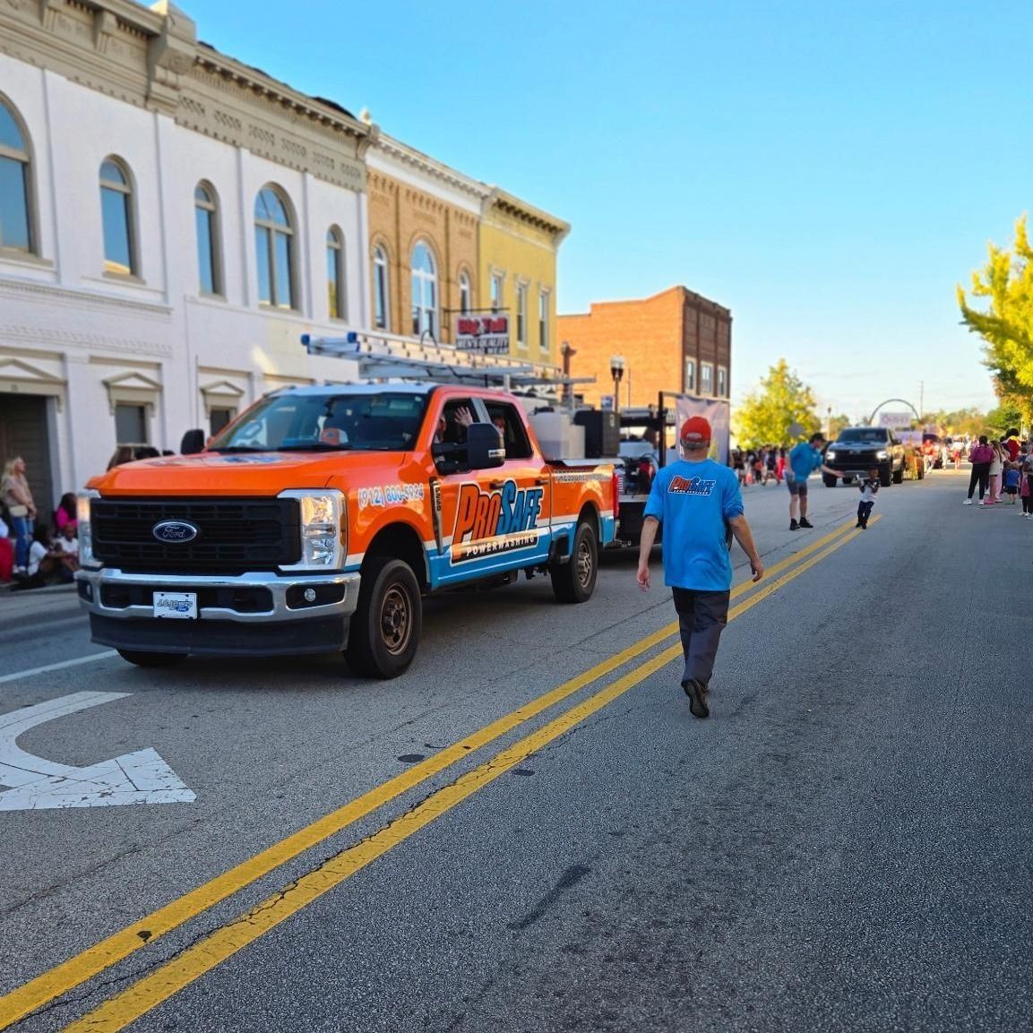 An orange and blue pickup truck drives down a city street during a parade, with people walking along the road.