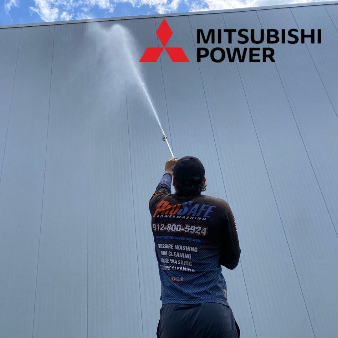 A technician uses a pressure washer on a gray building wall featuring the Mitsubishi Power logo.