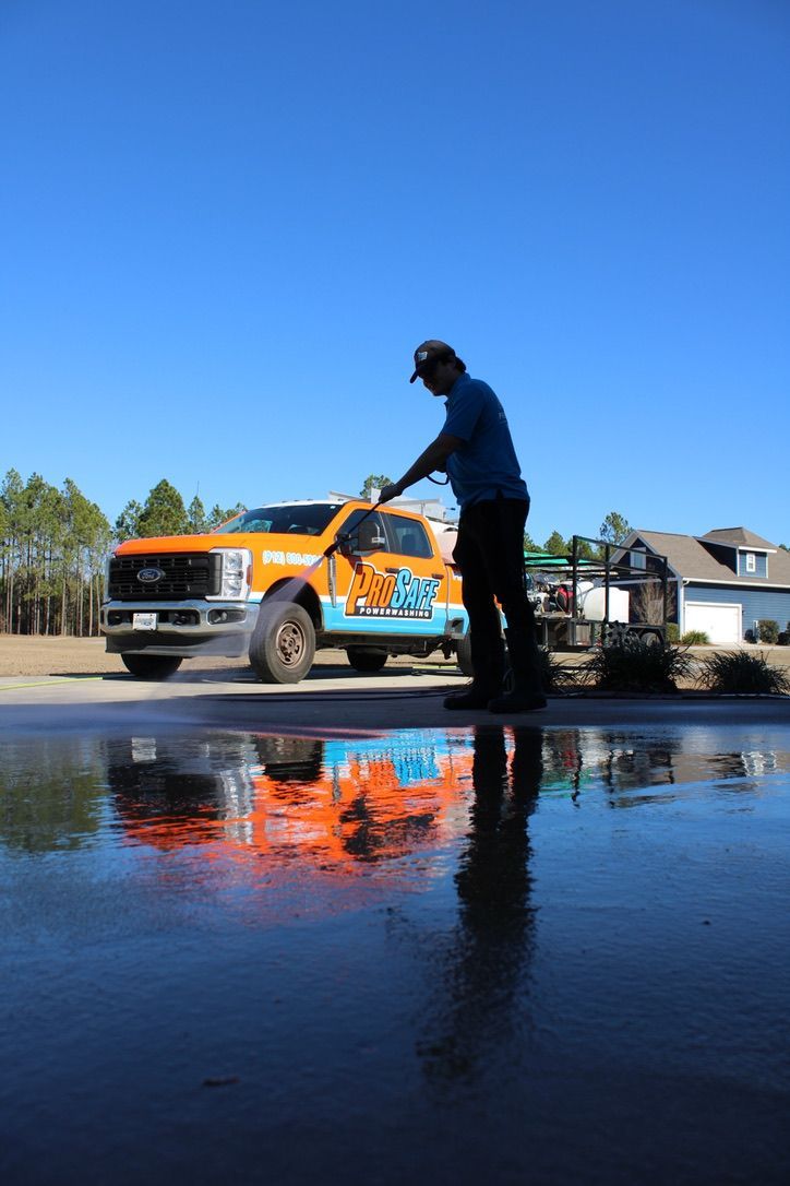 A worker power-washes a driveway, with their bright orange service truck reflected in the water-covered concrete.
