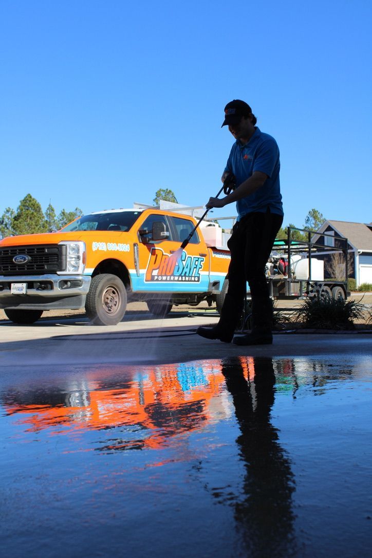 A person power washing concrete, with their orange and blue service truck visible nearby, reflected in a large puddle.