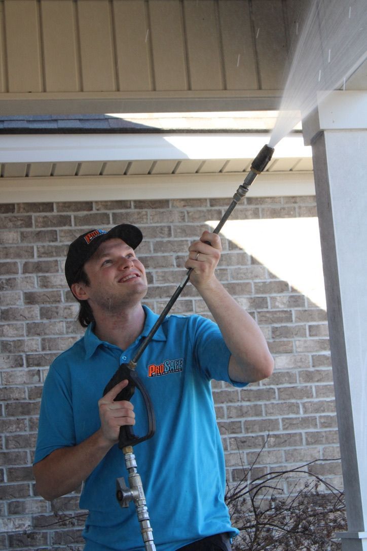 A person in a blue uniform power washes the underside of a porch overhang in front of a brick house.