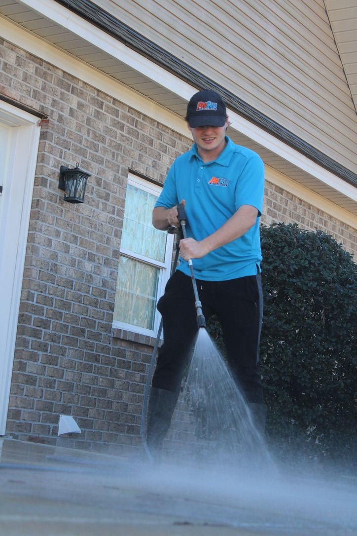 A person in a blue polo shirt and dark cap uses a pressure washer to clean a concrete driveway in front of a brick house.