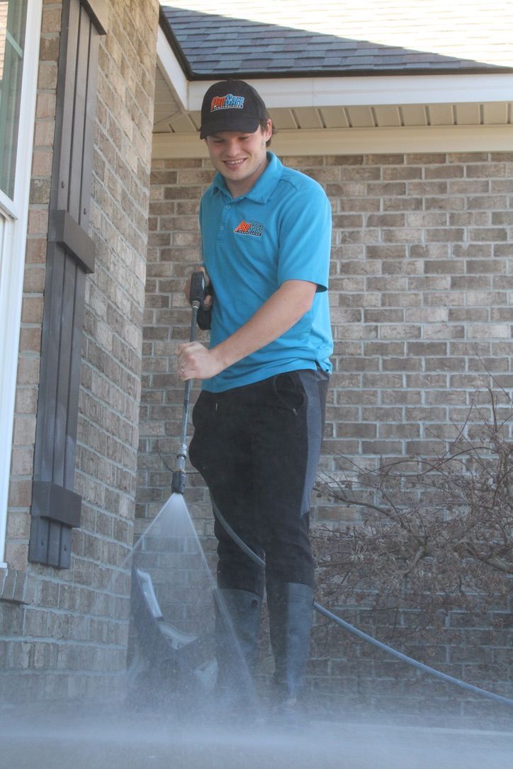 A person in a blue polo shirt and cap pressure washes a sidewalk next to a brick house.