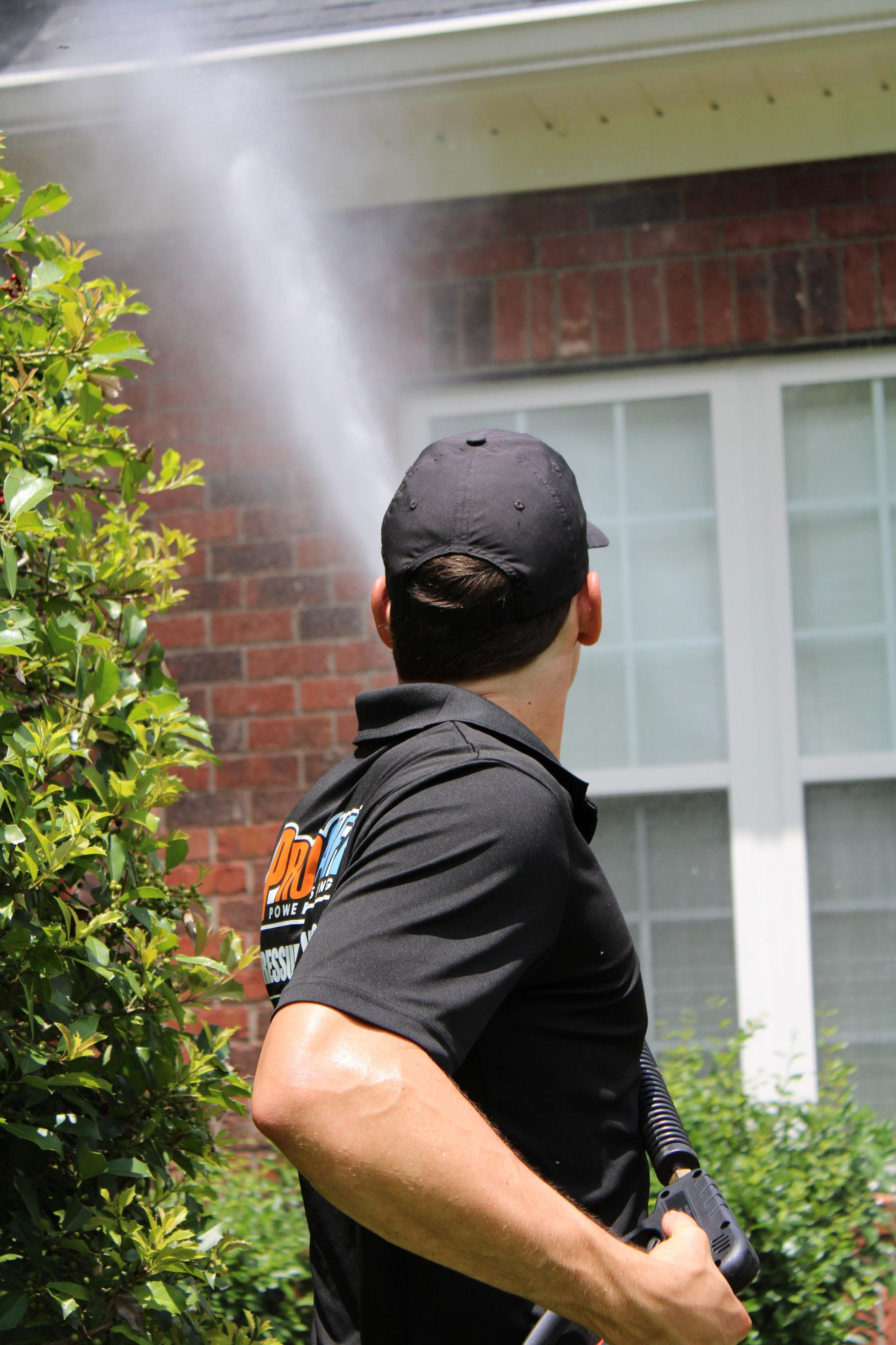 A person in a black uniform power washing the exterior brick wall of a house near a window.