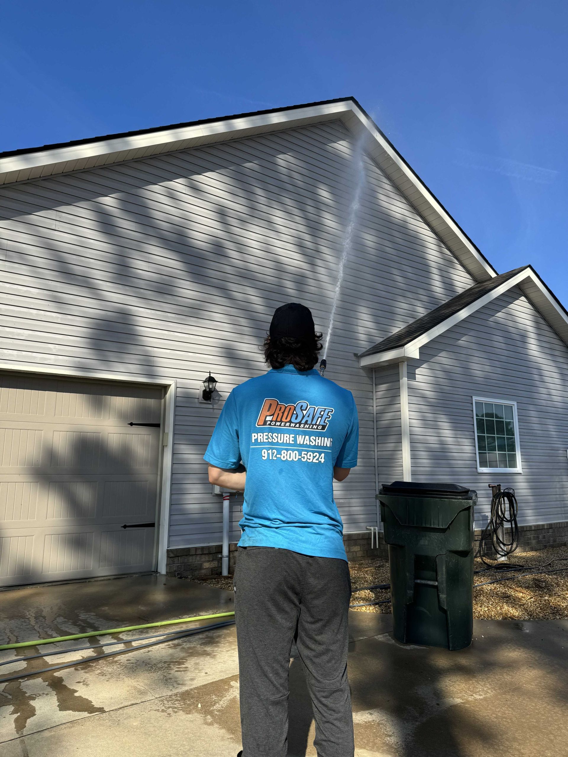A person in a blue shirt pressure washes the side of a white house on a sunny day.