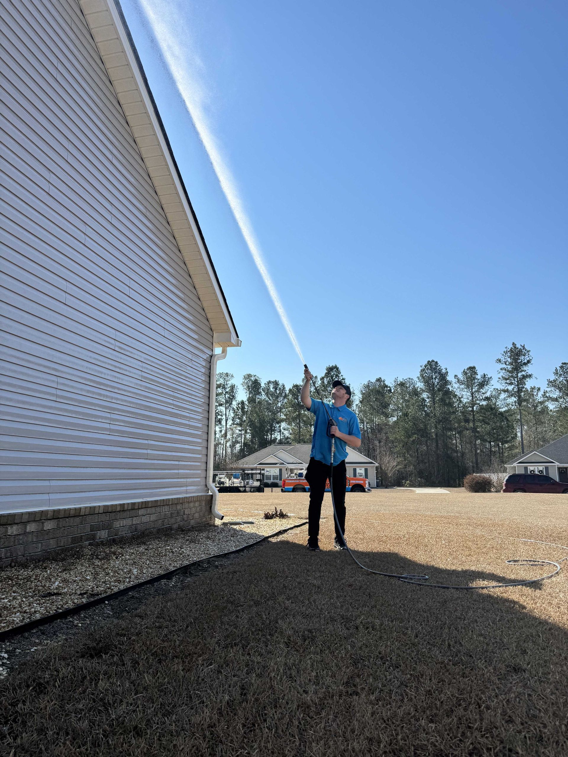 A person in a blue shirt uses a pressure washer to spray the siding of a light-colored house under a clear blue sky.