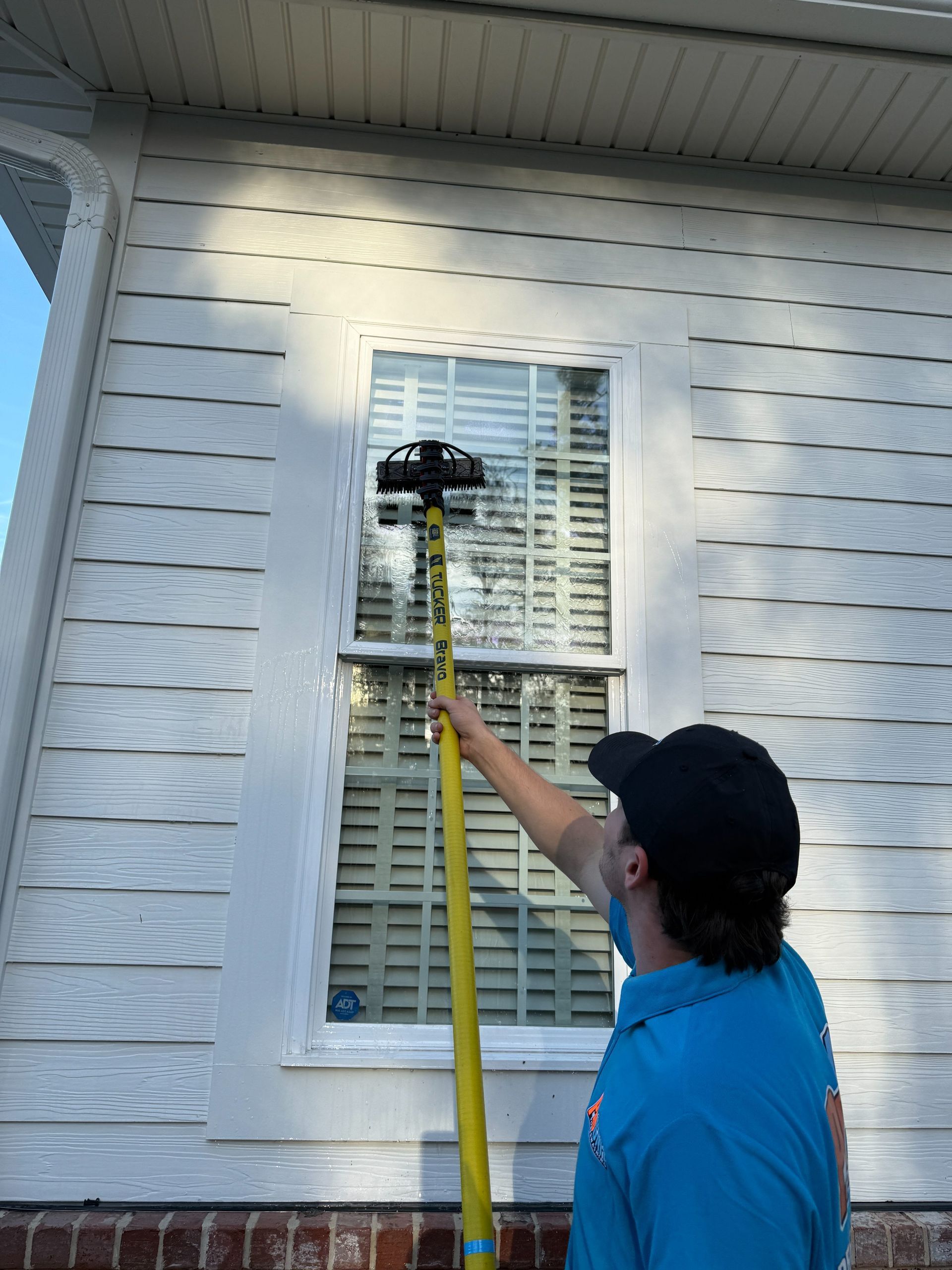 A person in a blue shirt uses a yellow pole with a brush attachment to clean an exterior house window.