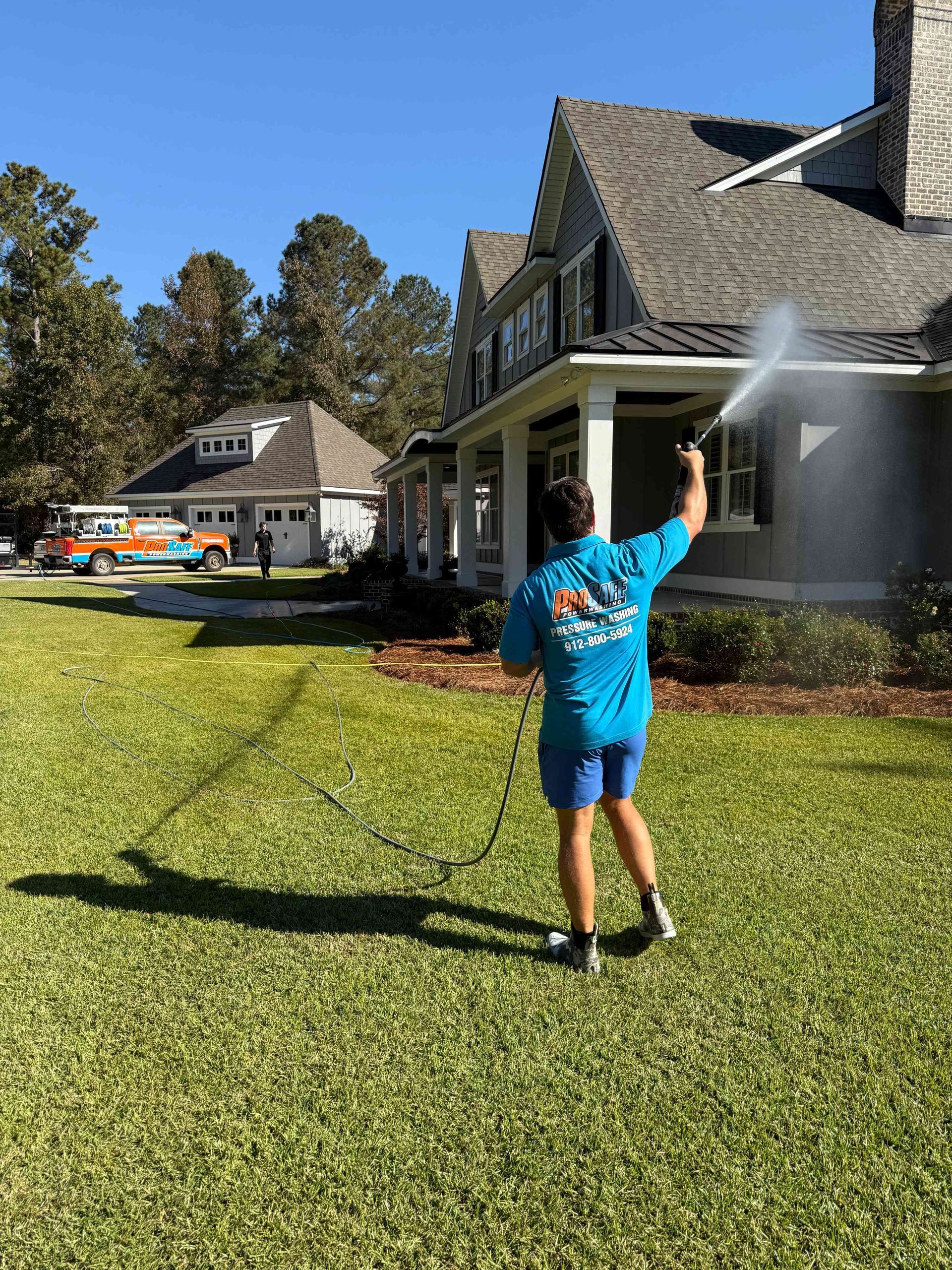 A worker in a bright blue uniform sprays the side of a large house with a pressure washer in a grassy yard.