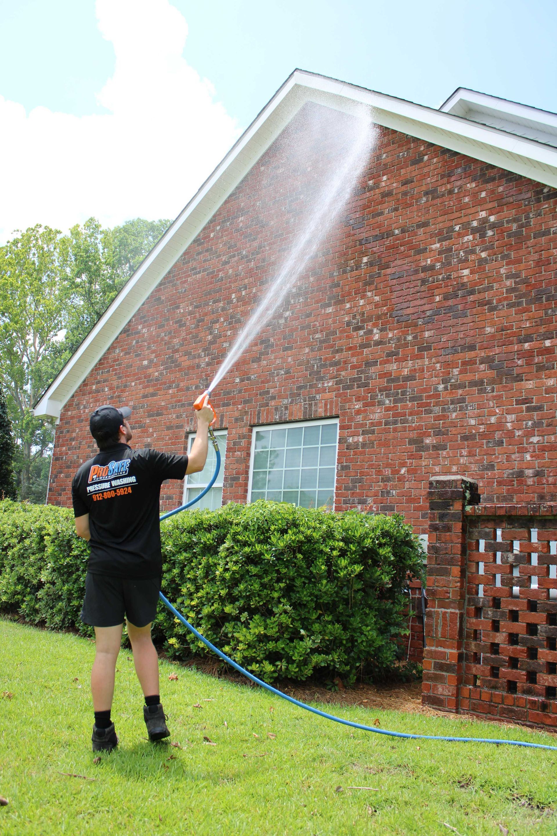 A person in a black uniform sprays water onto a brick wall while standing in a yard.