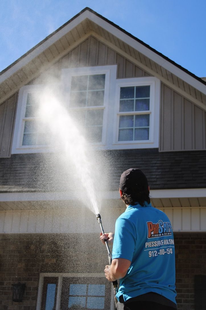 A worker in a blue uniform uses a pressure washer to clean the exterior of a two-story home.
