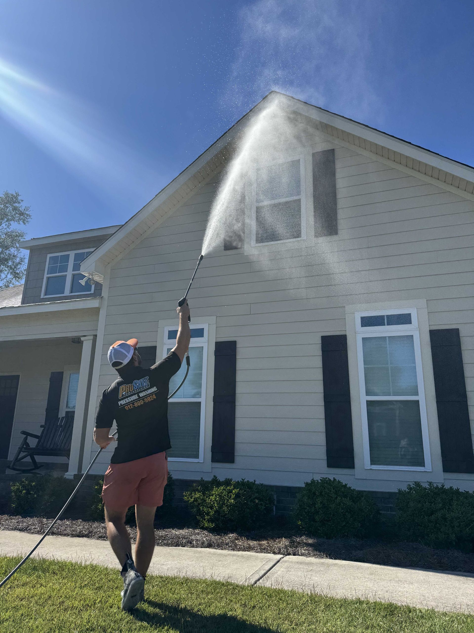 A person in a black shirt and shorts uses a pressure washer to clean the side of a light-colored house on a sunny day.