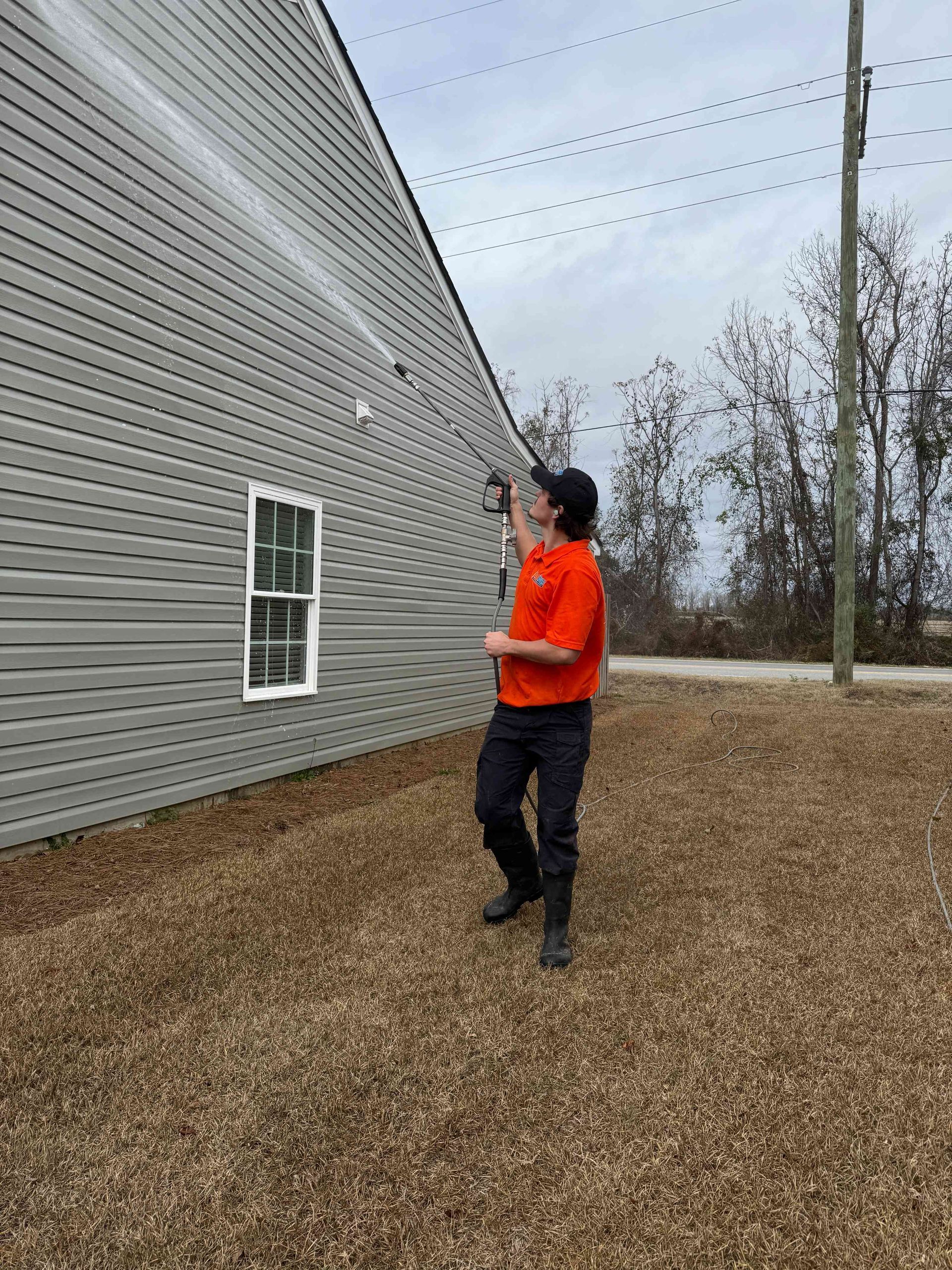 A technician in an orange shirt uses a long extension pole to power wash the light gray vinyl siding of a house.
