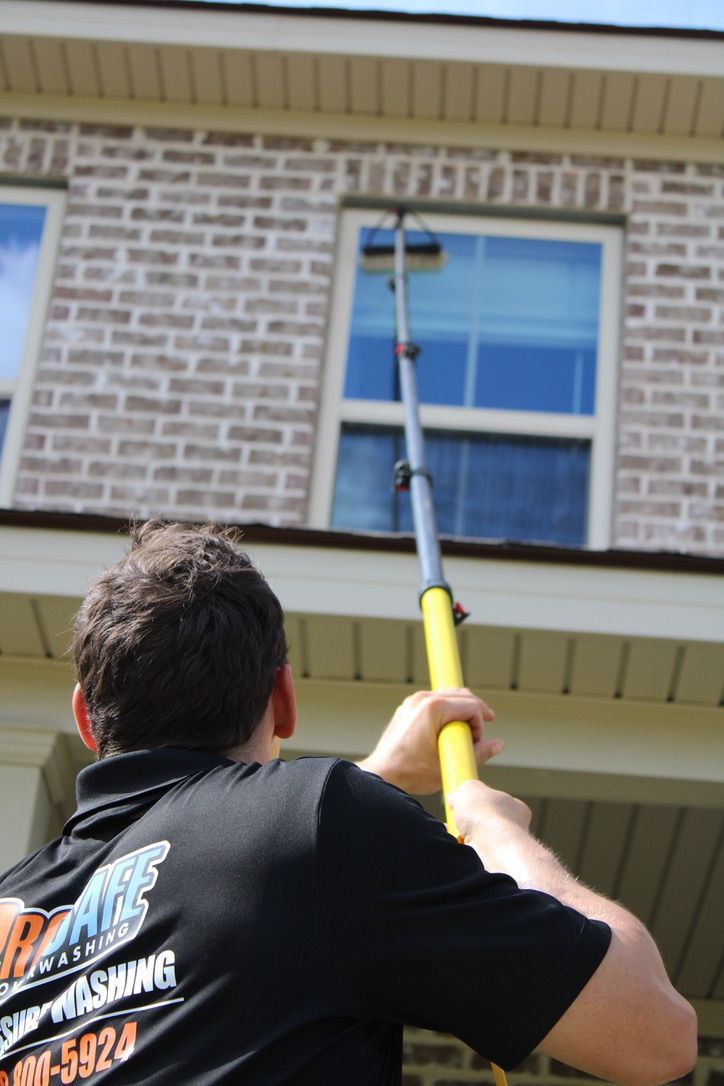 A window washer uses an extension pole to clean the exterior second-story window of a brick house.