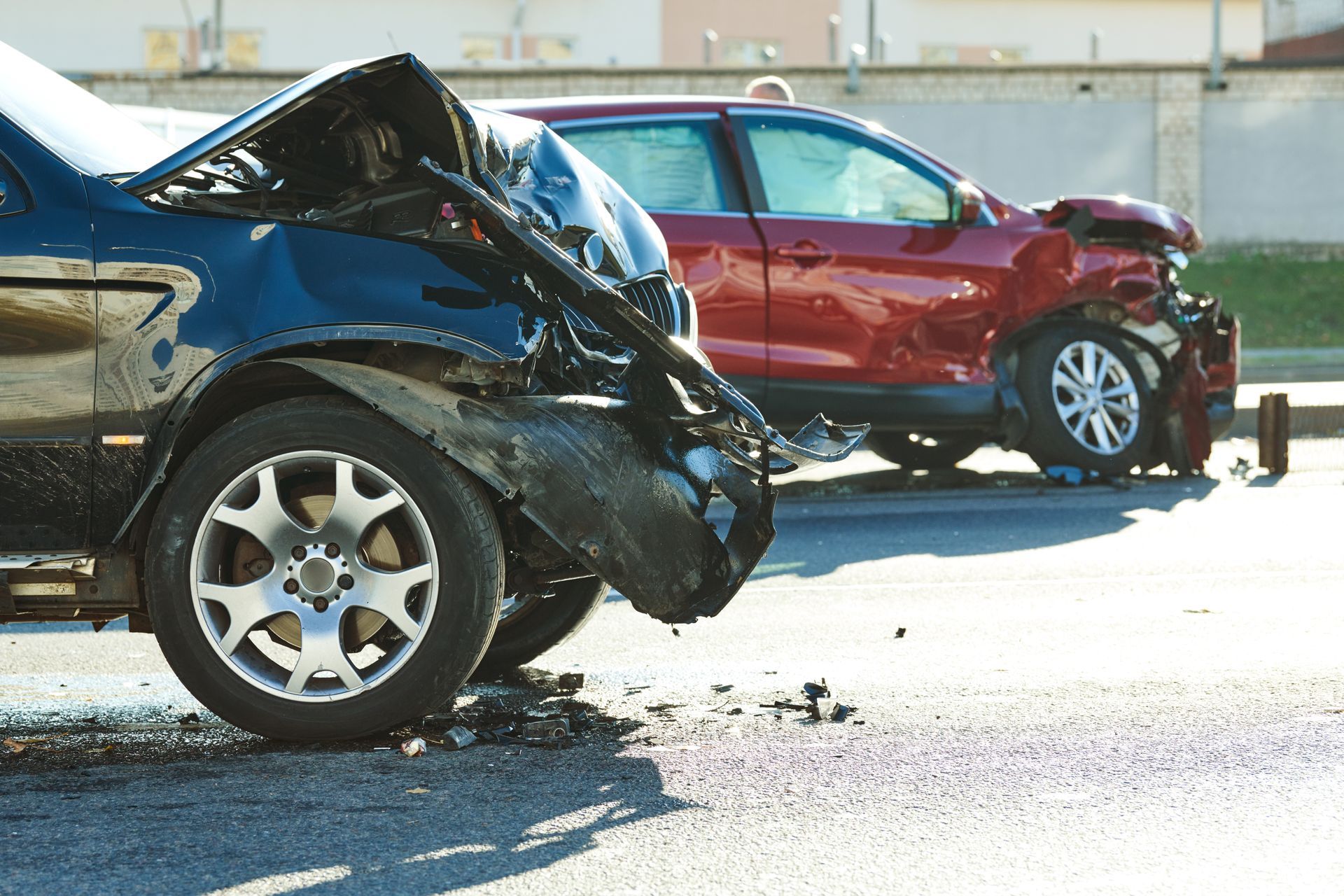 Two damaged cars stopped on a road after a collision, with crumpled fronts and debris nearby.