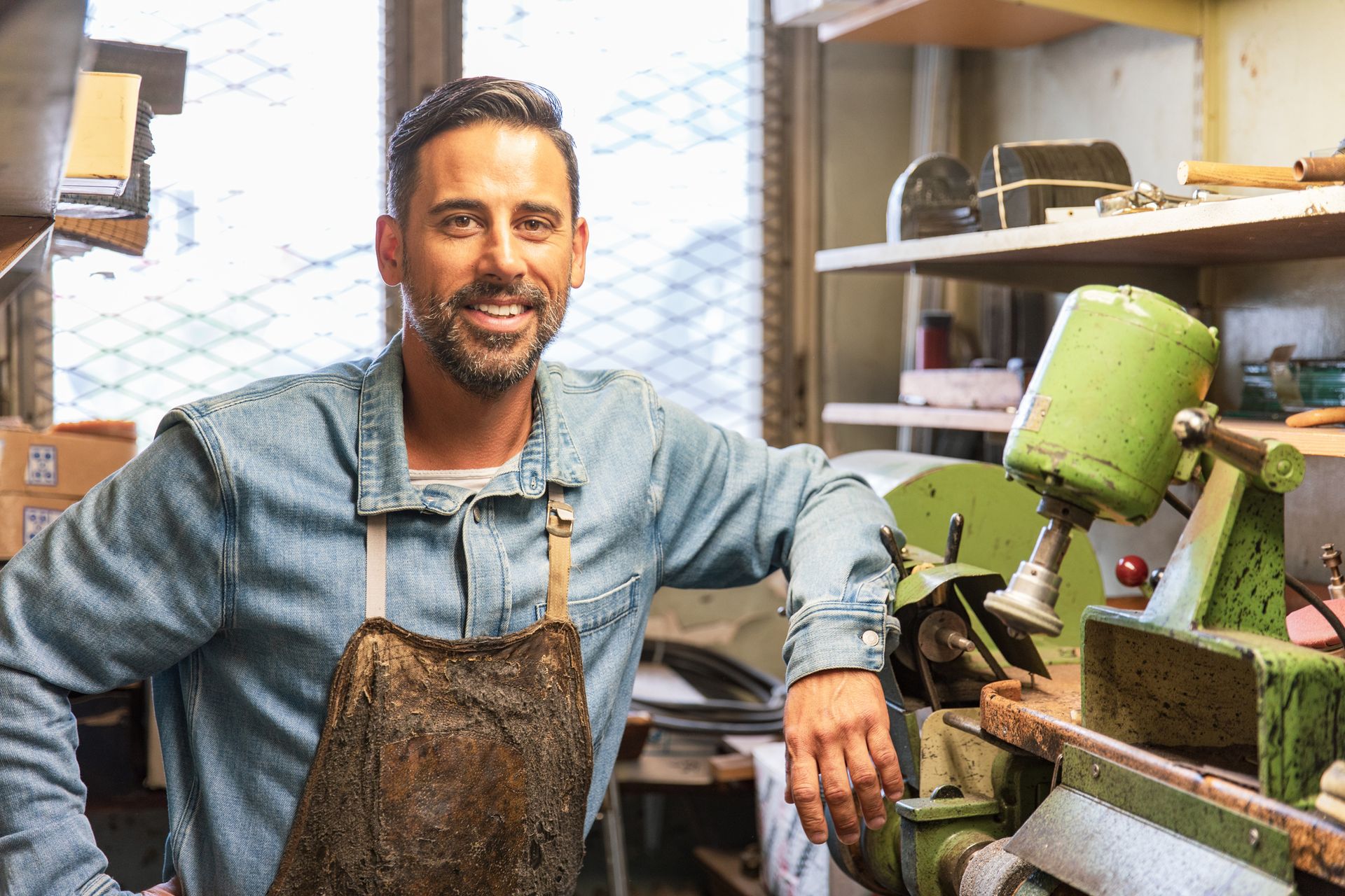 Man in workshop, smiling, wearing apron and leaning on machine.