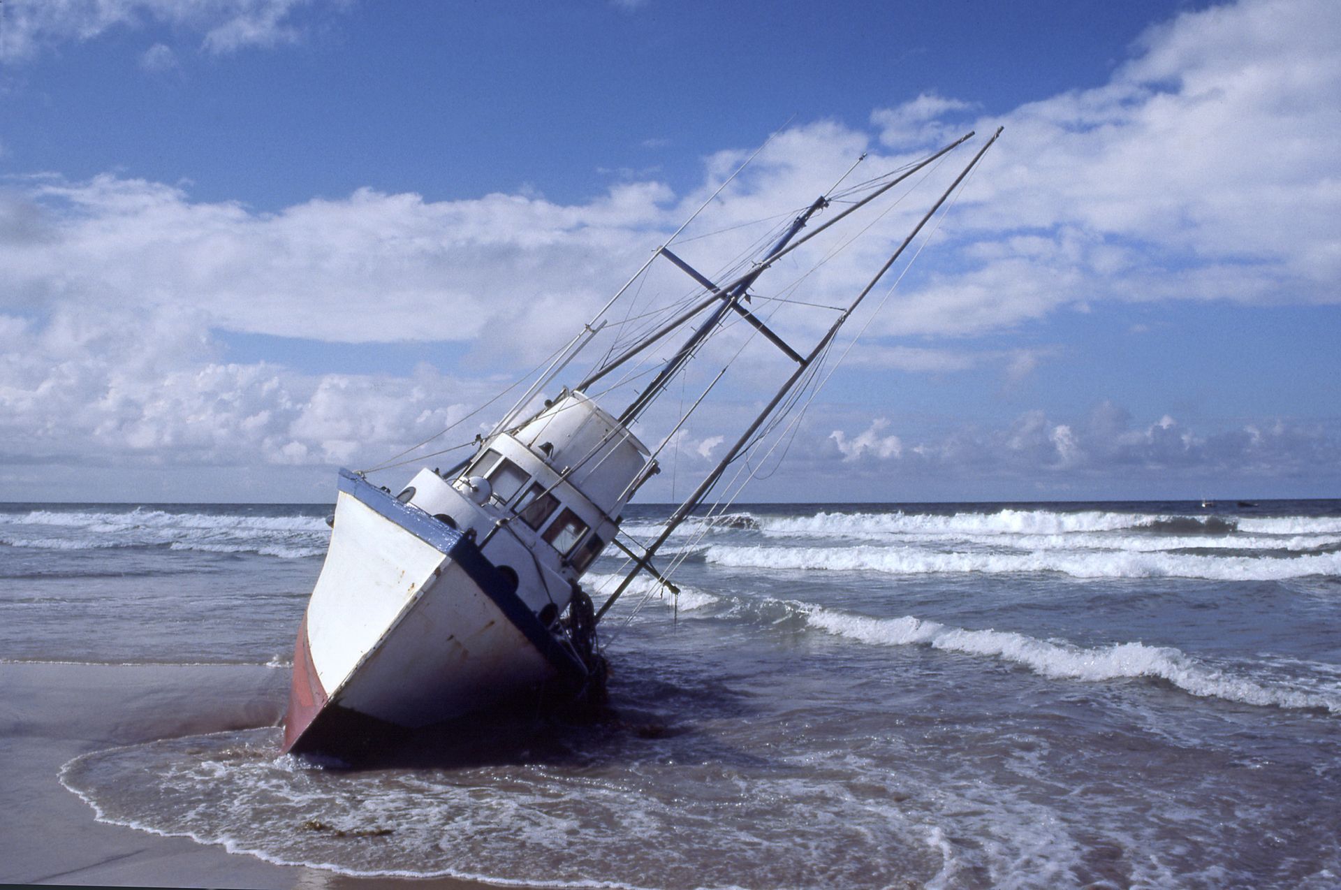 A stranded white and blue boat on a sandy beach, tilting toward the ocean.