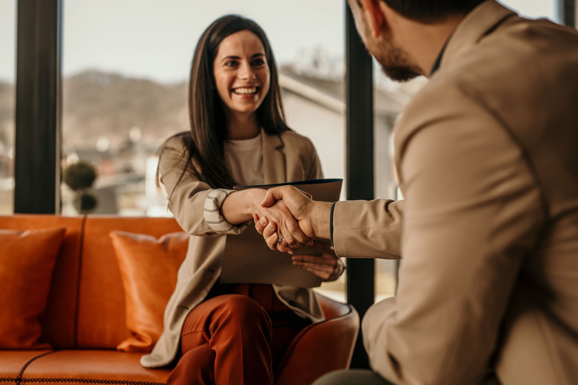 Woman in business attire shaking hands with a man, both smiling. Interior, orange sofa.