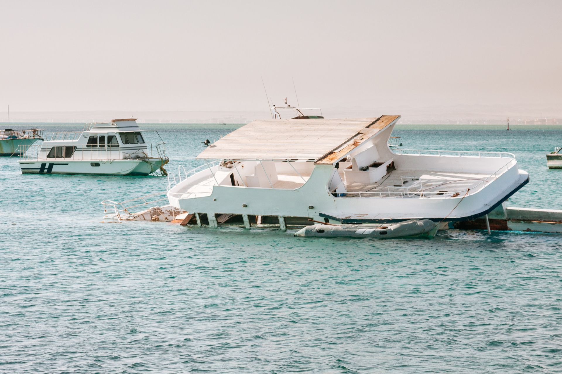 White yacht partially submerged in the water with damage, other boats in the background, sunny day.