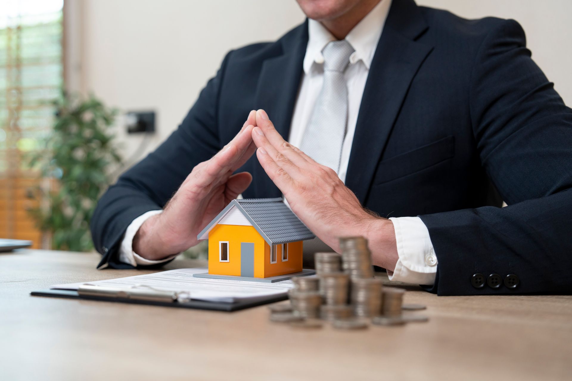 Man in suit protecting model house with hands, coins, and documents on table.