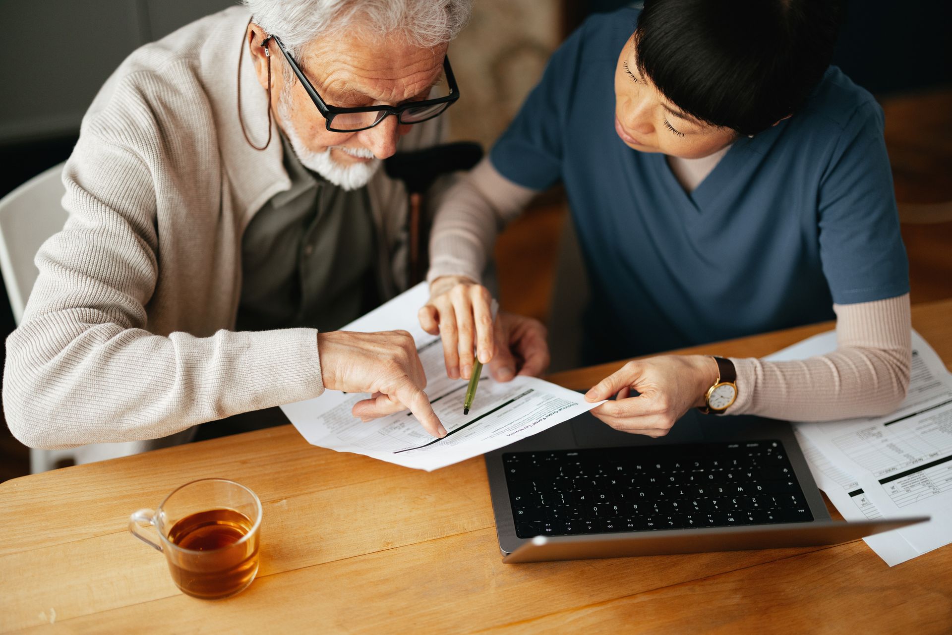 A senior man and a healthcare worker review papers at a table with a laptop and tea, indoors.