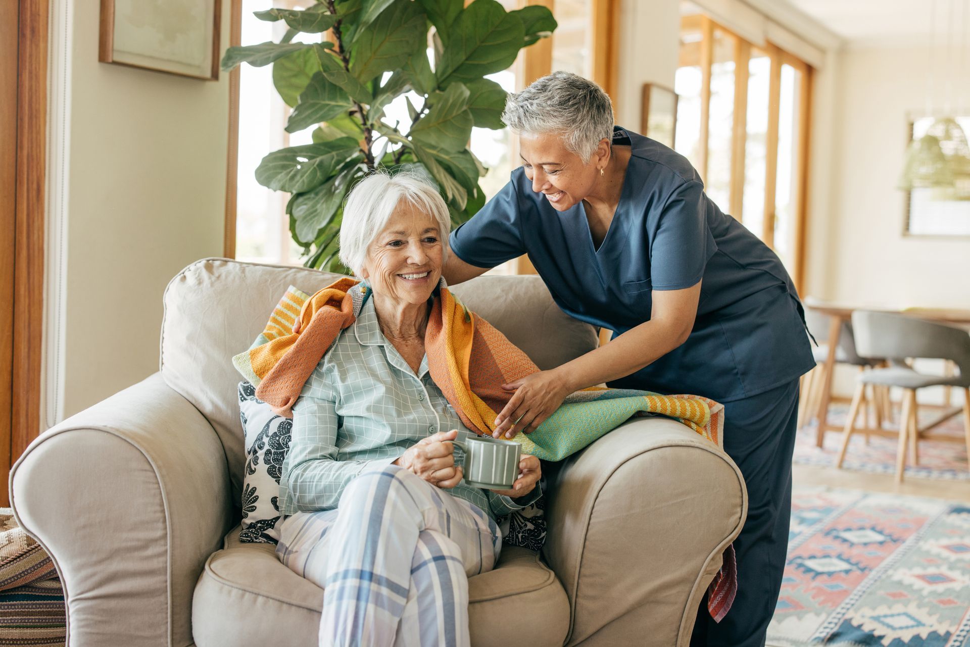 Caregiver placing a blanket on an elderly woman seated in a chair. Bright living room setting.