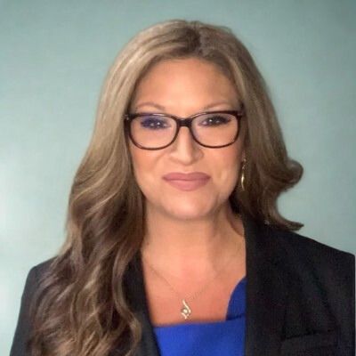 Woman wearing glasses and a black blazer, smiling at the camera. Turquoise background.